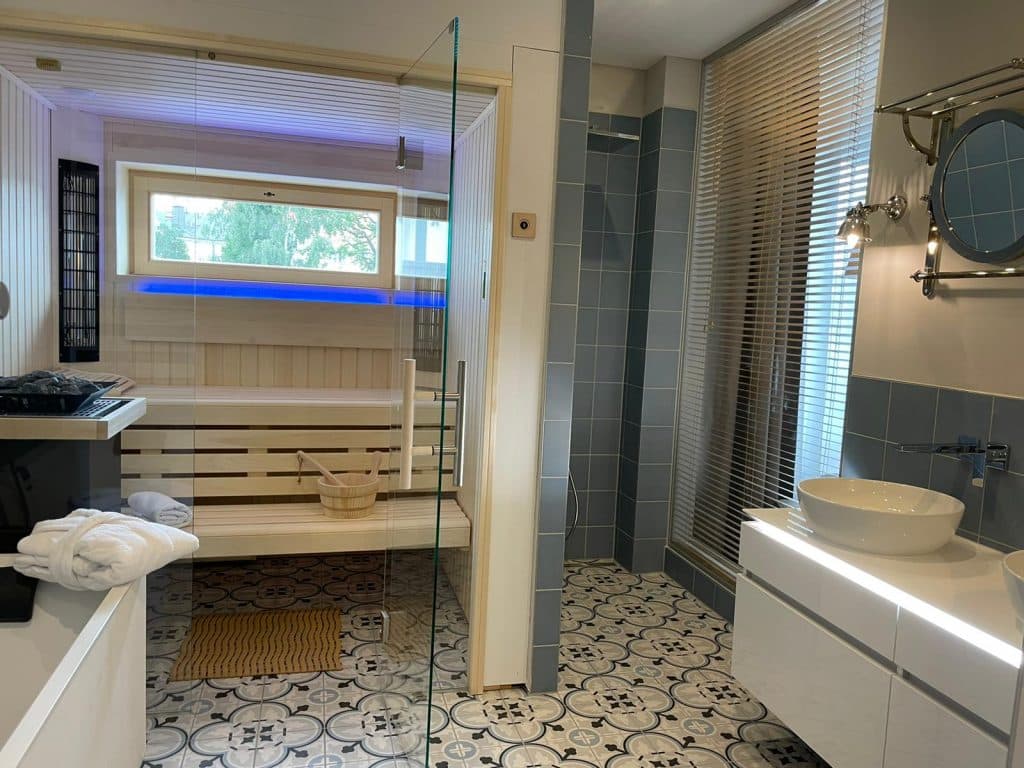Modernes Badezimmer mit gemustertem Fliesenboden, glasüberdachter Sauna mit Holzbänken, blauer Akzentbeleuchtung, Gefäßwaschbecken auf weißem Waschtisch, rundem Spiegel und hohem Fenster mit Jalousien, das Tageslicht hereinlässt.